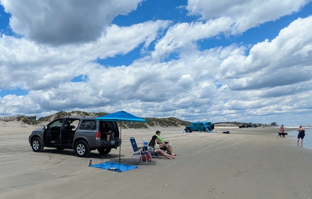 A black eight-passenger four-wheel-drive SUV parked on the sand with a shade canopy, towels and chairs set up beside it.