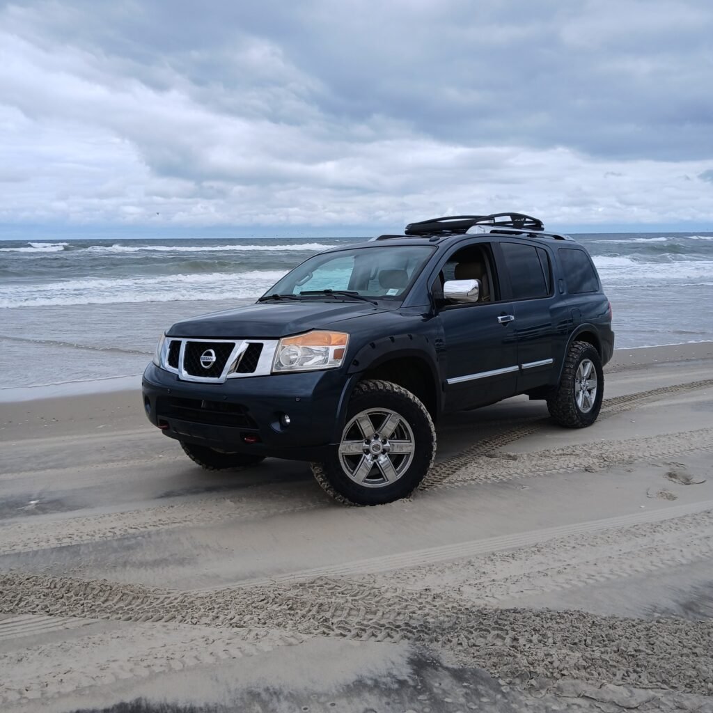 A black Nissan Armada four-wheel-drive SUV parked on the sand with the surf and an overcast sky behind it.