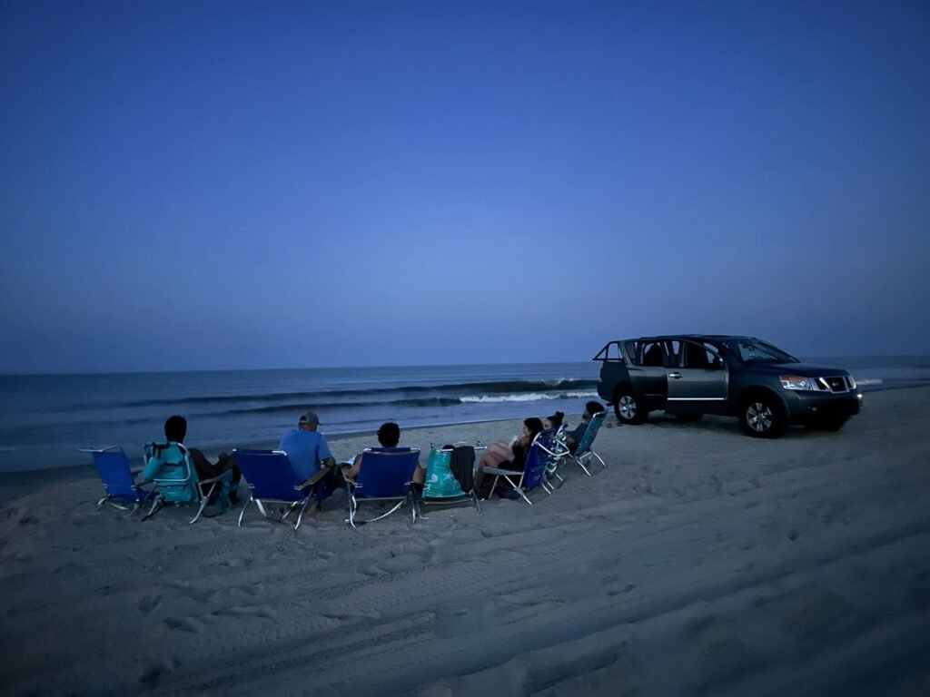 A group seated in beach chairs at blue hour on Carova beach with a black four-wheel-drive SUV parked alongside.