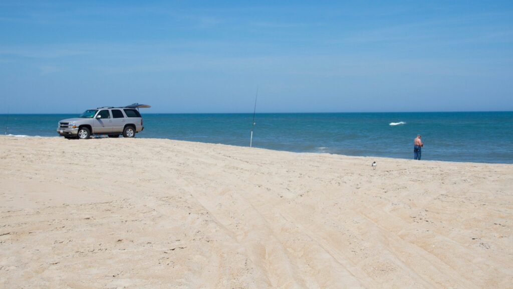 A white four-wheel-drive SUV parked on the open sand at Cape Hatteras National Seashore, ocean to the left and dunes behind.