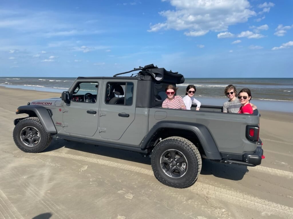 A grey Jeep Gladiator with its doors removed, parked on the beach with passengers riding in the back.