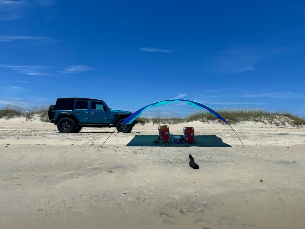 A rental Jeep parked on the sand beside a shade canopy with two people sitting underneath on a clear day.