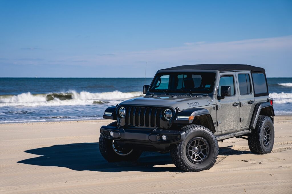 A grey four-door Jeep Wrangler parked on the sand with breaking surf behind it.