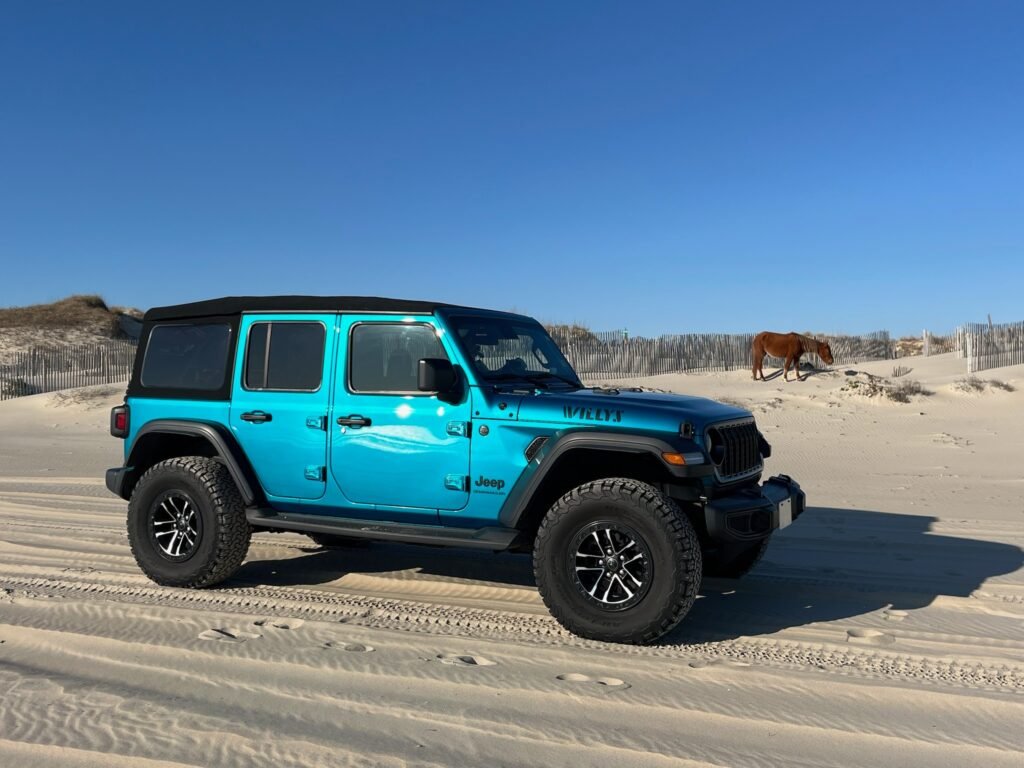 A turquoise four-door Jeep Wrangler parked on hard-packed sand with the dune line and open beach behind it.