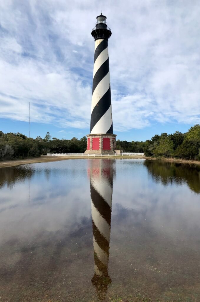 The black and white spiral-banded Cape Hatteras Lighthouse mirrored in standing floodwater at Buxton, framed by pines and a blue sky.