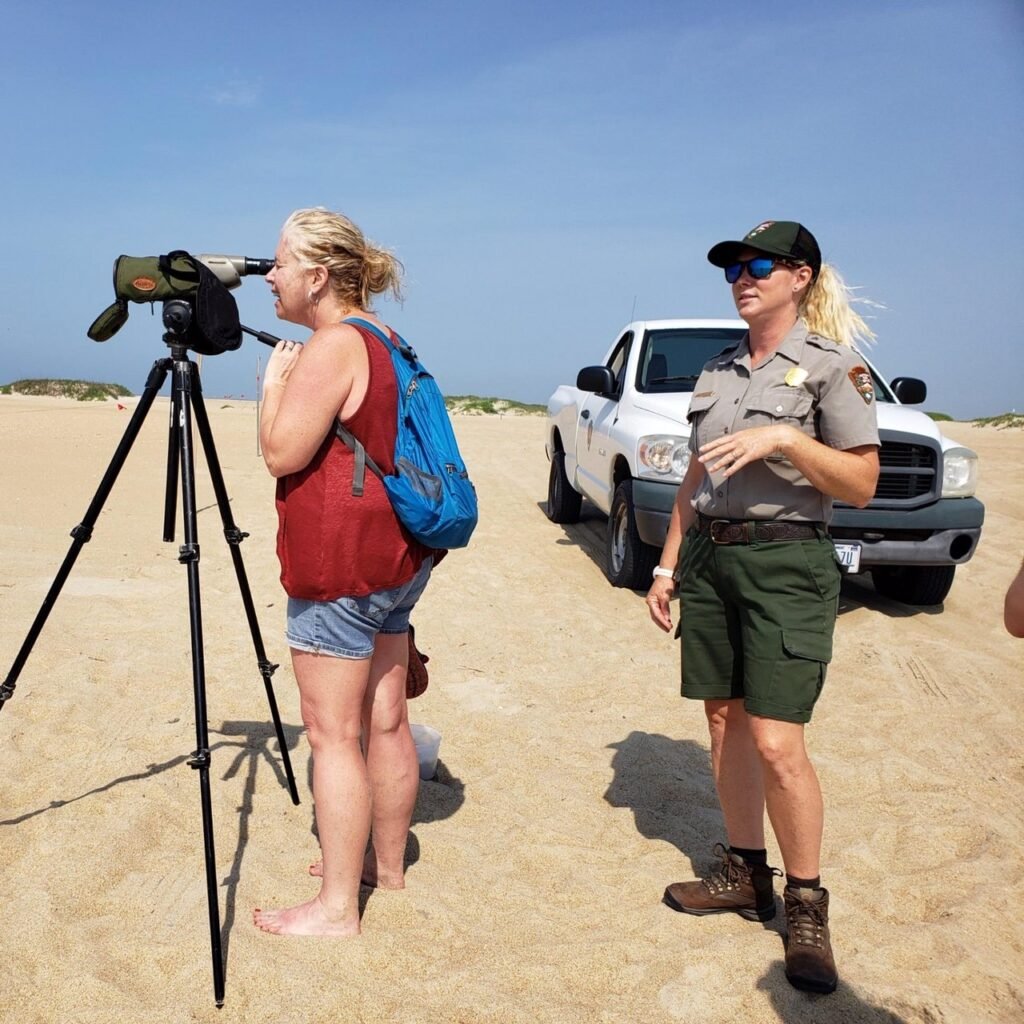 A National Park Service ranger talks with beach visitors gathered around a spotting scope on the sand at Cape Point, a pickup truck parked nearby.